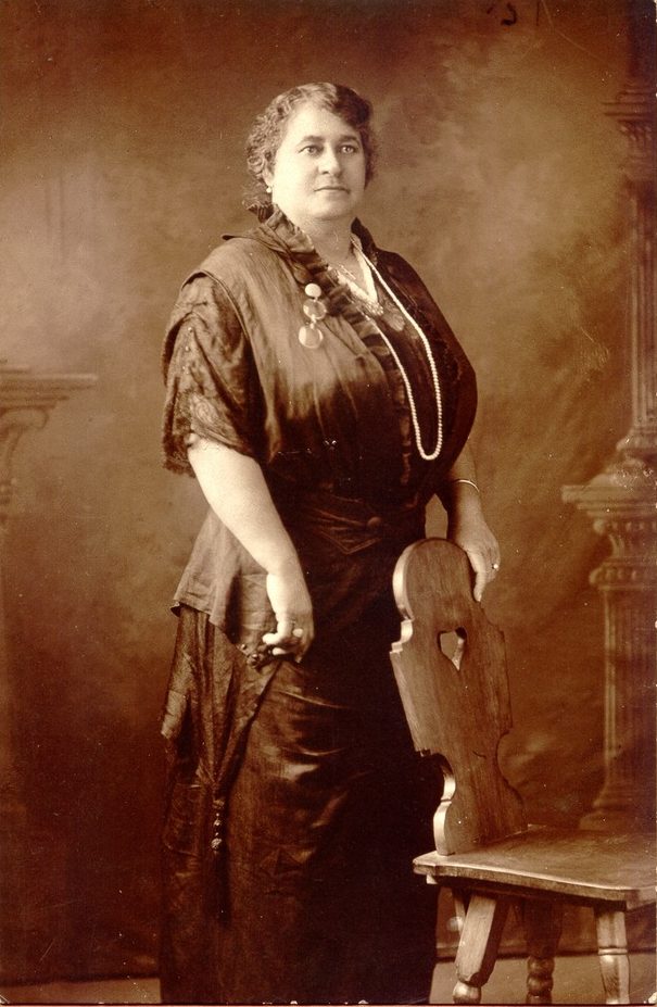 Portrait of Maggie Walker, the first Black woman bank president in the United States, who advocated for her community in Richmond, Virginia. Walker is standing with one hand resting on a chair. She wears a long dress made of dark fabric and has an assortment of jewelry. 