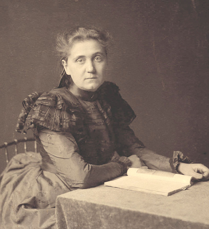 An image of Jane Addams seated at a table. She is facing the lens with her hands resting near an open book on the table. She is wearing a long dress. Her hair is pulled back. 