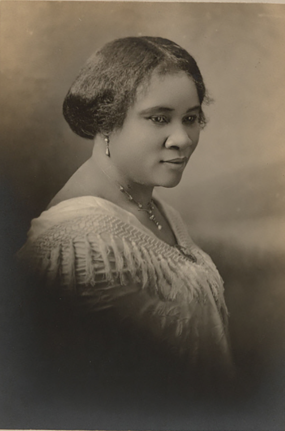 A photograph of Madam C.J. Walker, a self-made Black female millionaire. Her chest, shoulders, and head are visible. Her shoulders are covered by a draped piece of clothing. She wears a necklace and earrings. Her hair is pulled back. 