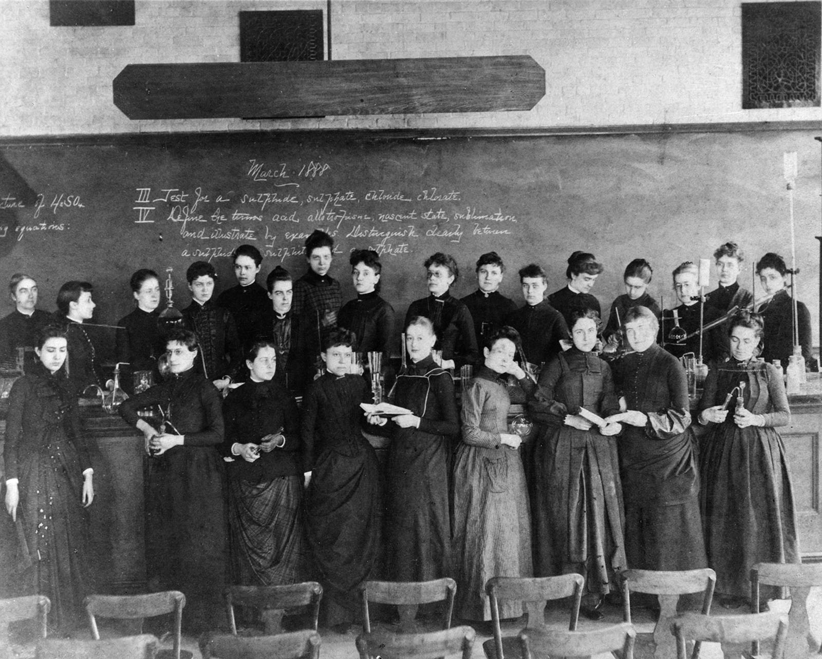 Ellen Swallow Richards and female college students in two lines in front of a classroom. Richards is the figure all the way on the left in the back row. All of the students with her are wearing dark dresses. They are all white. The chalkboard behind them indicates that the date is March 1888. 