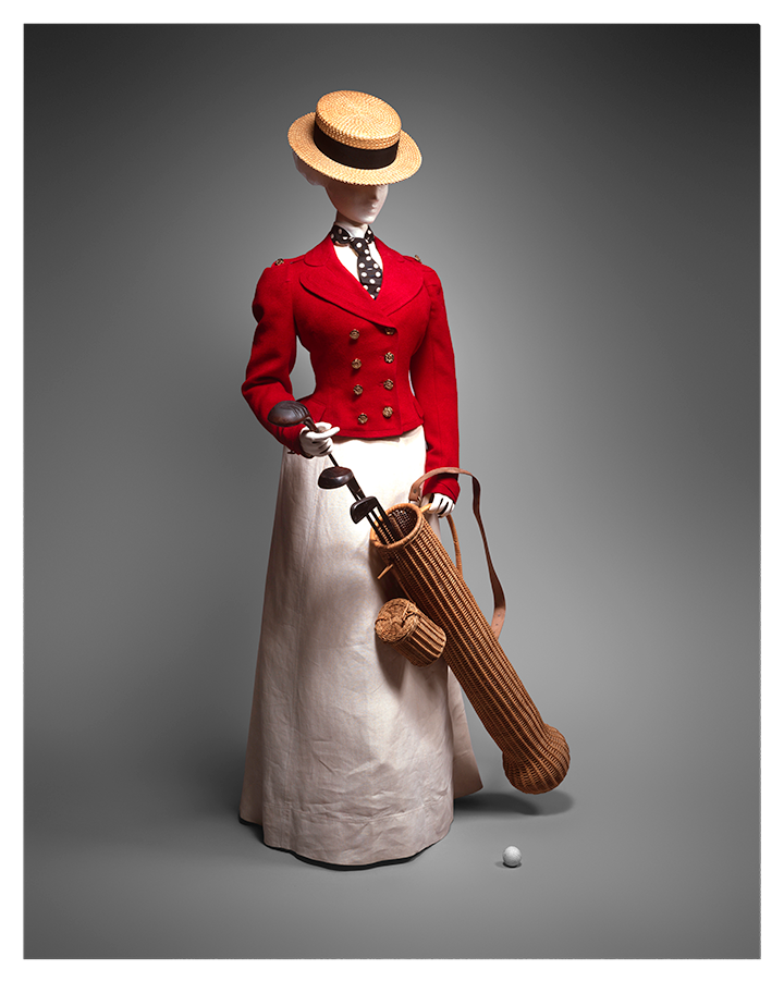 A women’s golf outfit from the 1890s with a round tan hat, fitted red button-down jacket, and a long white skirt. A polka dot tie is tucked into the jacket. The mannequin holds a golf bag made of what appears to be wicker.