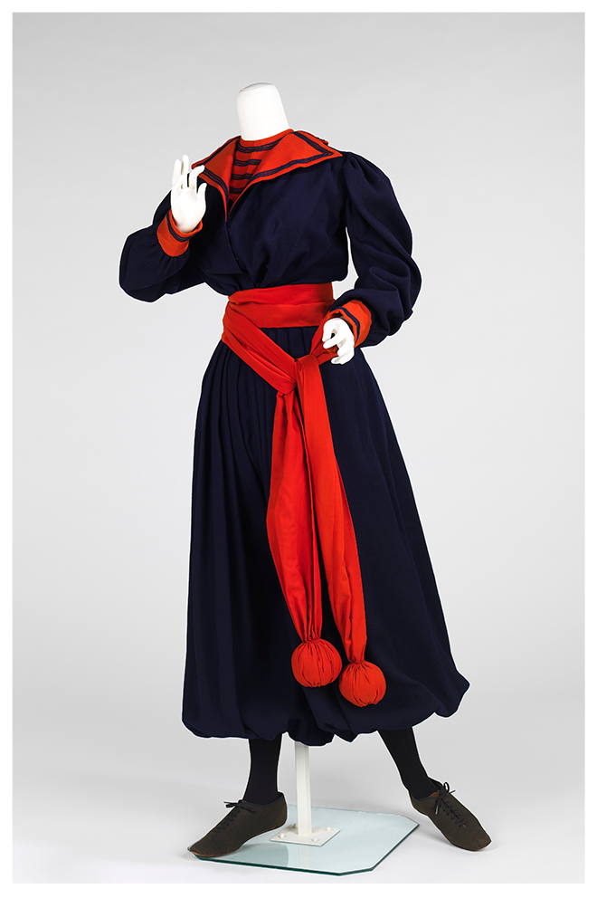 A gym suit from the 1890s with a baggy top in dark fabric and cropped pants that looks like a skirt and balloon out from the waist. The collar and cuffs, are red. A long piece of red fabric that looks like a sash is tied around the mannequin’s waist and has weighted balls on the end. These would have been used for exercise activities.