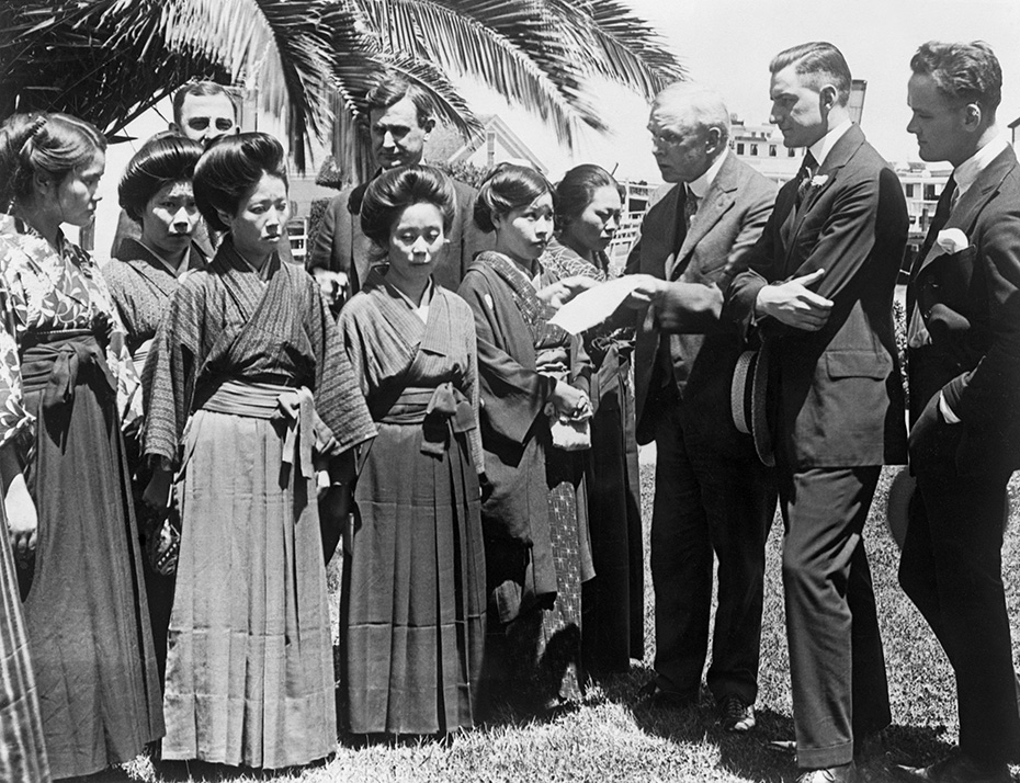 Black and white photograph of Japanese women in traditional kimonos (left) and white government officials (surrounding the women) reviewing their passports. The image was taken at Angel Island in 1920.
