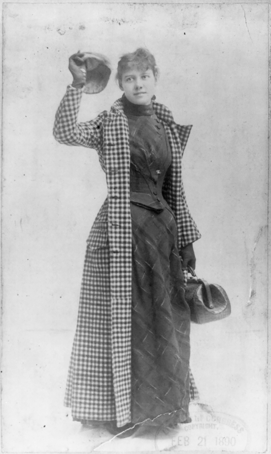 An image of Nellie Bly taken around 1890. She is standing holding a cap in her right hand and a bag in her left. She is wearing a long, dark, dress and a checkered coat. Her hair is pulled back. 