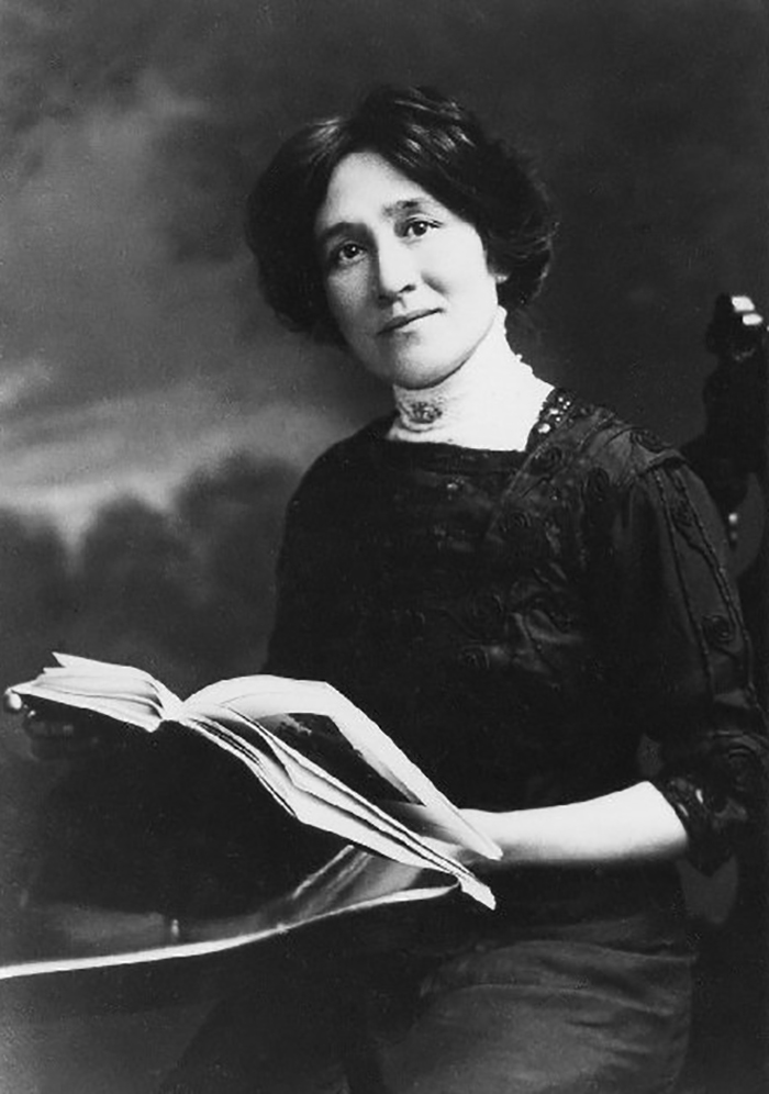 An image of Edith Maude Eaton holding a book. She is seated at a table and wearing a long, dark, dress. 