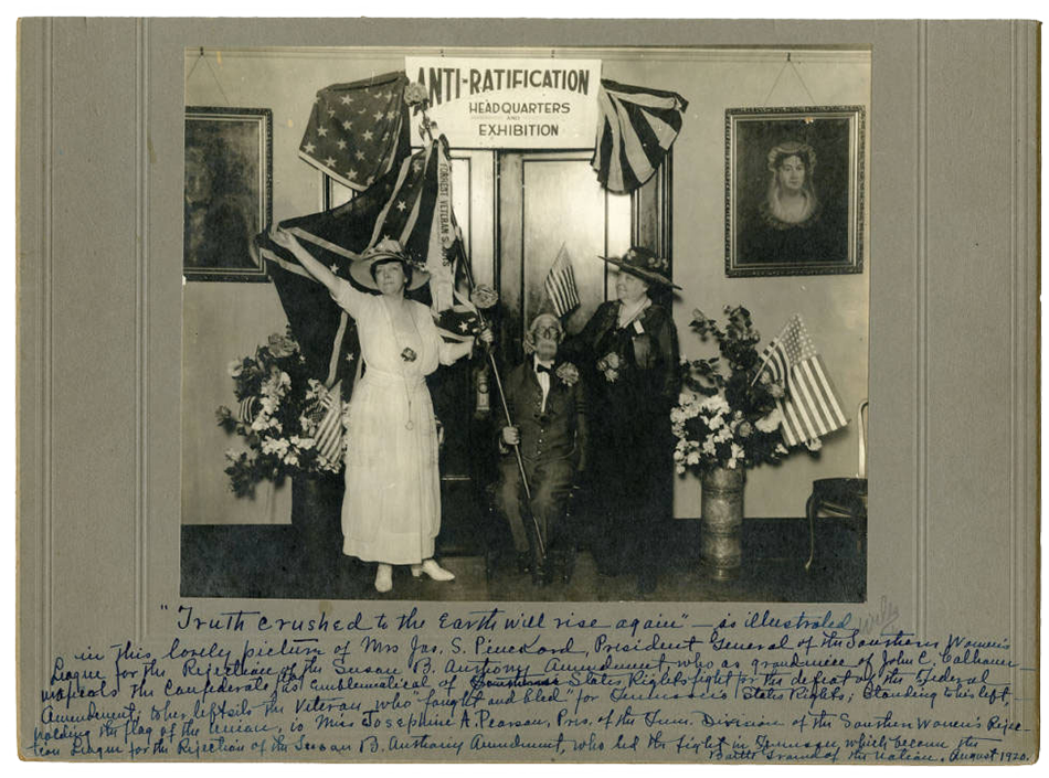 Black and white photograph taken at the anti-suffrage headquarters. A group of three white people pose for the picture. The figure on the left is a woman in a white dress holding a Confederate flag. In the middle, sits an old man in a suit who is also holding the Confederate flag. The figure on the right is a woman in black who appears to be holding a small American flag. Behind the women are two portraits and flower vases with both flowers and American flags inside them. The text: “Anti-Ratification Headquarters Exhibition” reads the banner hung above them. Below the photograph is a handwritten description of the photograph that is transcribed in the caption below.