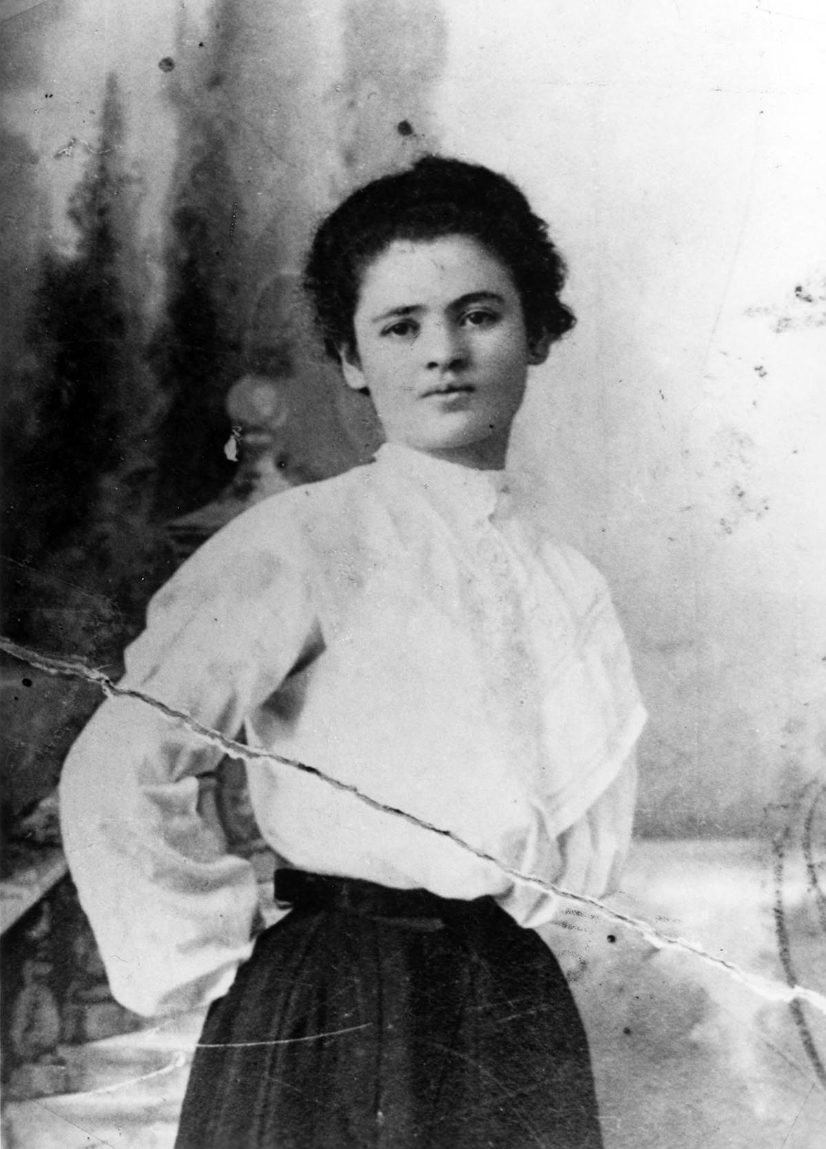 A portrait of Clara Lemlich in her mid-20s. She is standing with her hands behind her back, wearing a light colored blouse and dark colored skirt. Her hair is pulled back. 