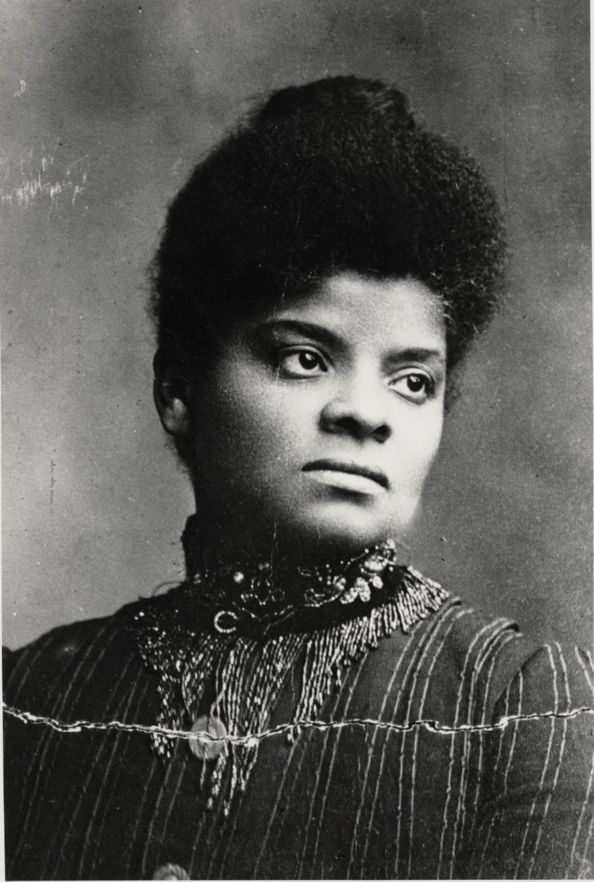 An image of Ida B. Wells around the age of 30. She is depicted from chest-level to the top of her head and wearing a dark top that covers her shoulders and neck. It has an elaborate collar. Her head is turned to the side and her hair is pulled back. 