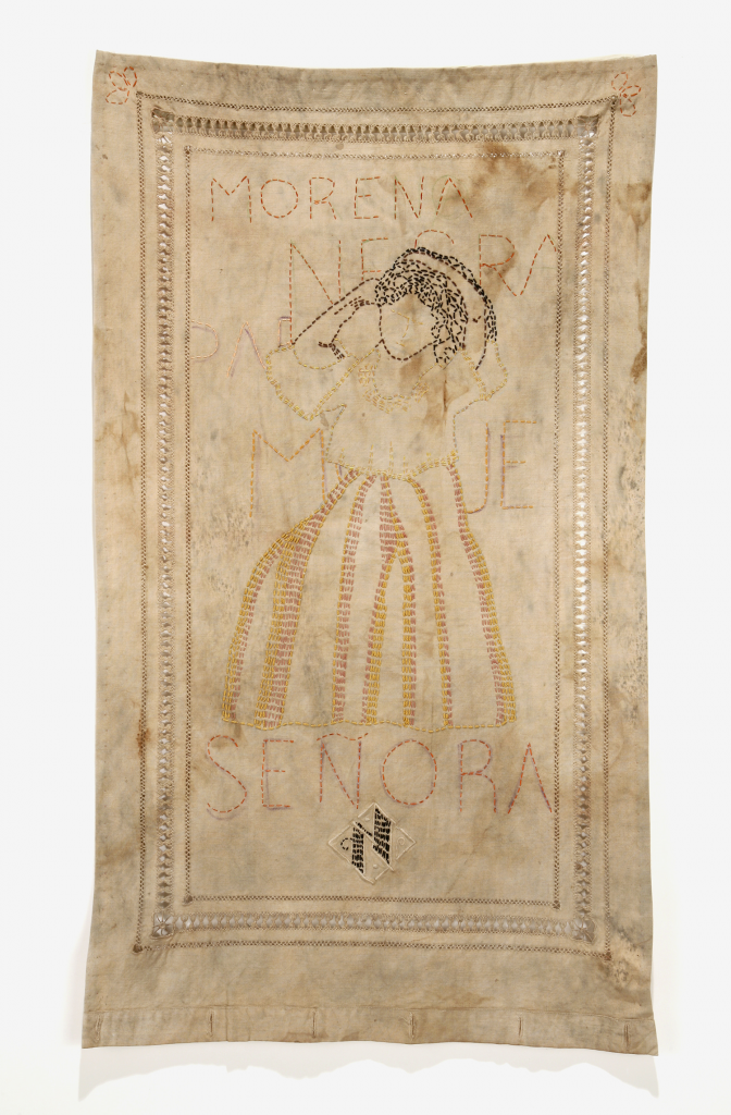 A piece of embroidery work, rectangular and stained, outlining the form of a faceless woman with dark hair. Above the figure, stitched in red thread appear the words “MORENA NEGRA” and “SENORA” below it. Dimensions: 30” (h) x 18” (w). 