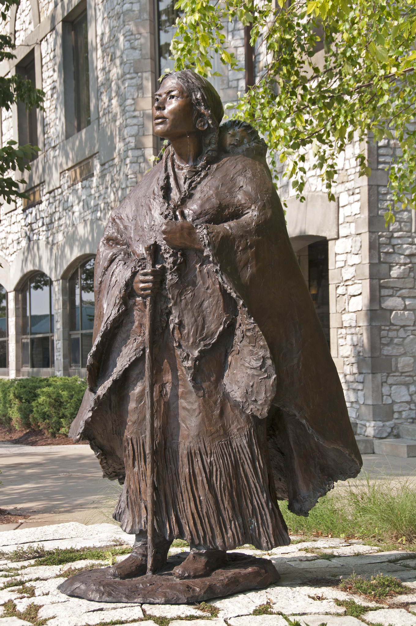 A 7 ft. tall bronze sculpture of a Lemhi Shoshone woman, Sacagawea, in Indigenous attire, standing with a walking stick and a child strapped to her back.